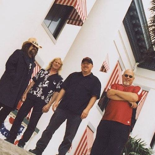 Four men are standing in front of a white building with red-and-white striped awnings. They are posing for the photo outdoors.