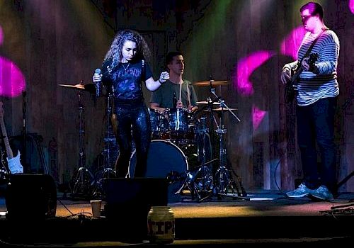 A live band performs on stage, with a vocalist, drummer, and guitarist under colorful lights and a dark background, creating a lively ambiance.