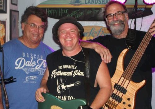 Three men standing together with musical instruments, smiling at the camera. They appear to be in a casual indoor setting, possibly at an event.