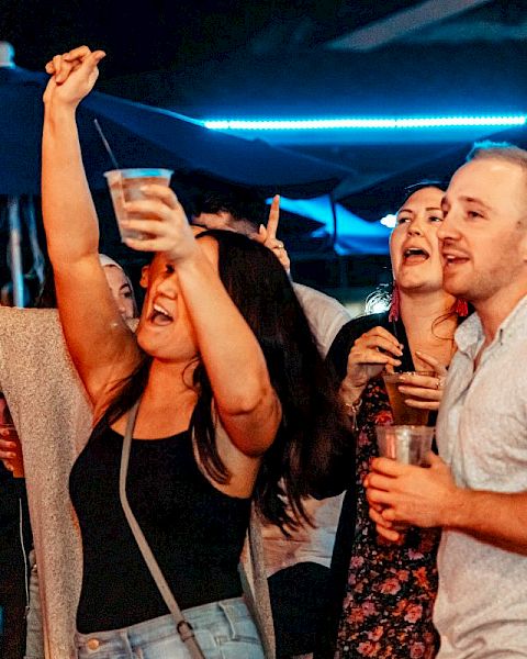 A group of people are enjoying themselves at a lively night event, holding drinks and cheering enthusiastically.