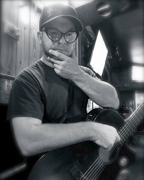 A man wearing glasses and a cap, holding their chin while their other hand rests on a guitar, in a dimly lit indoor setting.