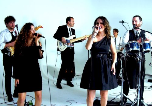 A band is performing with two vocalists, guitarists, a drummer, a keyboardist, and a percussionist in a studio setting, all dressed formally.