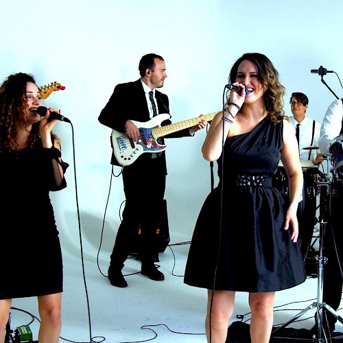 A band is performing with two vocalists, guitarists, a drummer, a keyboardist, and a percussionist in a studio setting, all dressed formally.