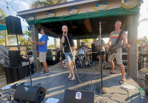 A band is performing live on an outdoor stage, featuring a singer, guitarist, bassist, and drummer. The setting looks like a casual beachside venue.