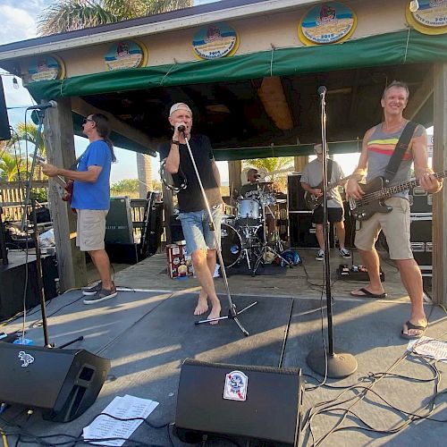 A band is performing live on an outdoor stage, featuring a singer, guitarist, bassist, and drummer. The setting looks like a casual beachside venue.