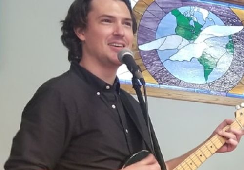 A person is singing and playing an electric guitar in front of a microphone, with a stained glass window in the background depicting a dove.