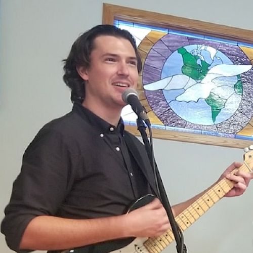 A person is singing and playing an electric guitar in front of a microphone, with a stained glass window in the background depicting a dove.