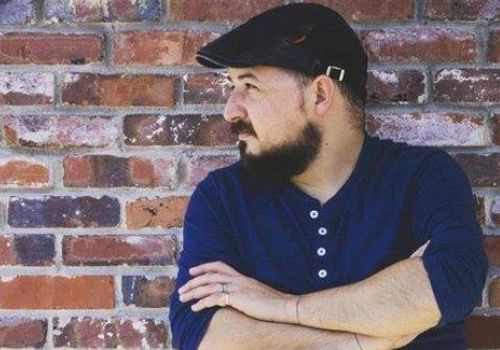 A man with a beard, wearing a flat cap and blue shirt, stands with crossed arms against a brick wall.