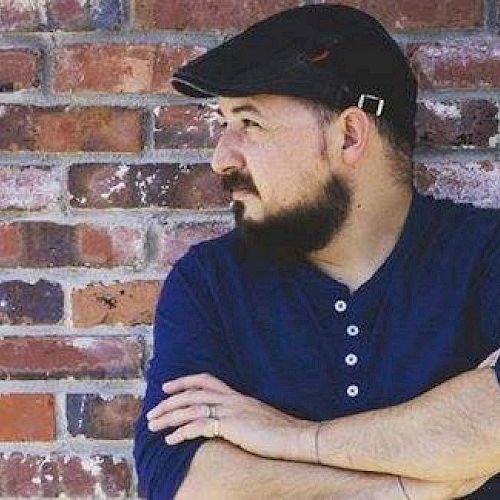 A man with a beard, wearing a flat cap and blue shirt, stands with crossed arms against a brick wall.