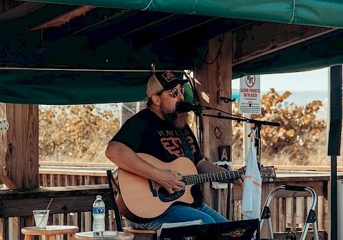 A musician with a guitar performs under a canopy, with stools, a drink, and a microphone stand nearby, in an outdoor setting.