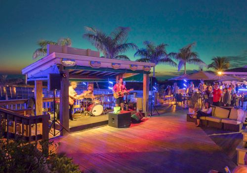 Outdoor night party with live band, colorful lights, people dancing and mingling on a wooden deck under palm trees.