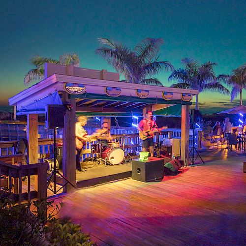 Outdoor night party with live band, colorful lights, people dancing and mingling on a wooden deck under palm trees.