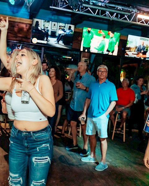 A group of people enjoying a lively bar or club, with one person taking a selfie and others watching televisions and drinking.