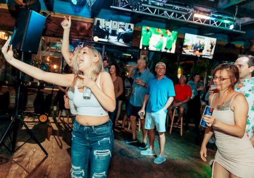 A group of people enjoying a lively bar or club, with one person taking a selfie and others watching televisions and drinking.