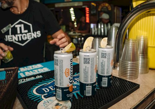 Cans with lemon slices on a bar mat, plastic cups beside them, and a person in a branded shirt nearby in a dimly lit bar setting.