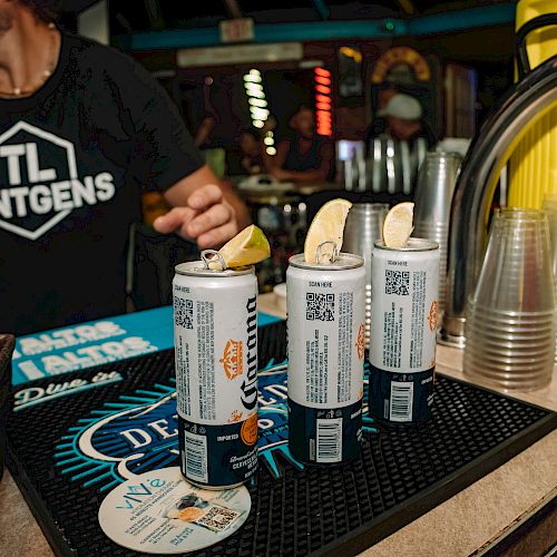 Cans with lemon slices on a bar mat, plastic cups beside them, and a person in a branded shirt nearby in a dimly lit bar setting.