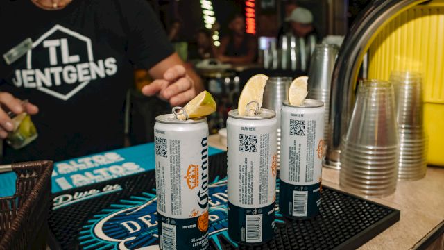 Cans with lemon slices on a bar mat, plastic cups beside them, and a person in a branded shirt nearby in a dimly lit bar setting.