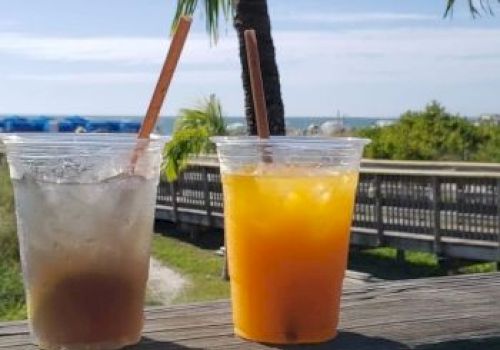 Two plastic cups with drinks, ice, and straws on a wooden railing; tropical beach background with a palm tree and boardwalk.