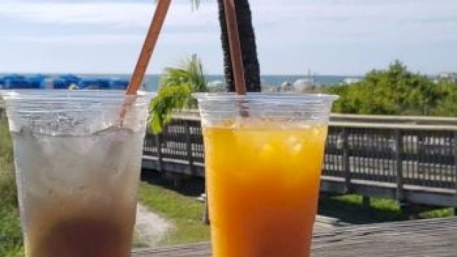 Two plastic cups with drinks, ice, and straws on a wooden railing; tropical beach background with a palm tree and boardwalk.