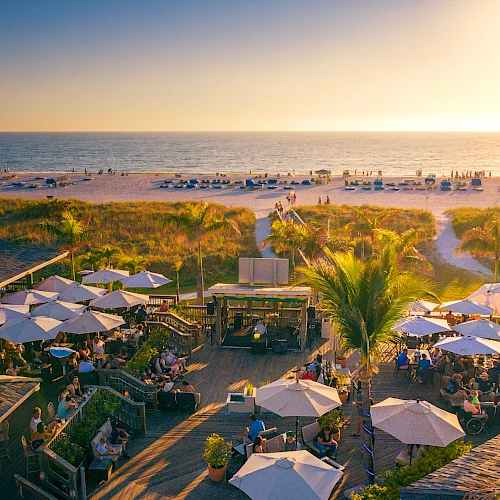 An outdoor beachside restaurant with many people dining under white umbrellas, overlooking the sea and sunset. Palm trees add to the tropical atmosphere.