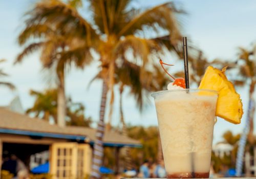 A tropical cocktail with a cherry, umbrella, and pineapple slice is set against a background of palm trees and a beachside bar.