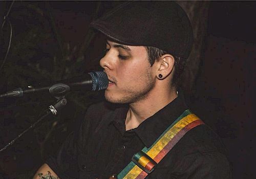 A person is playing a guitar and singing into a microphone, wearing a dark shirt and a cap, with a colorful strap on the guitar, and has a visible tattoo.