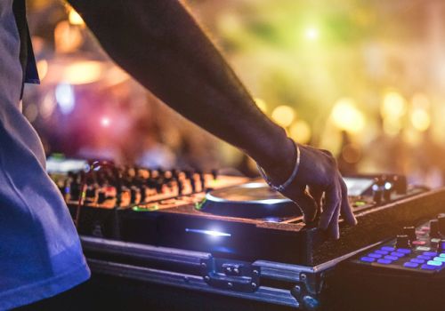 A DJ is mixing music on a turntable with colorful, blurred lights and a crowd in the background, creating a lively party atmosphere.