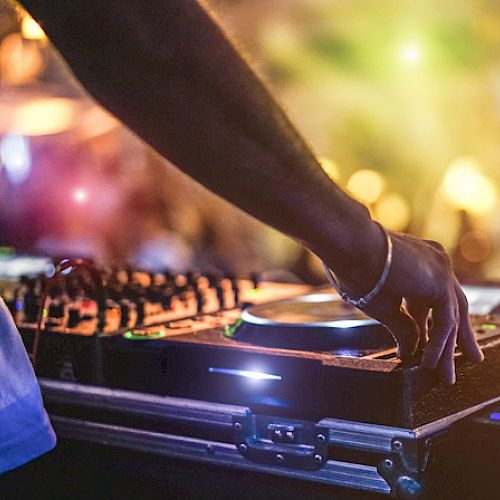 A DJ is mixing music on a turntable with colorful, blurred lights and a crowd in the background, creating a lively party atmosphere.