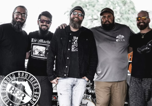 Five bearded men in sunglasses pose outdoors with a large &ldquo;BULL&rdquo; motorcycle-style logo, looking like a rock/biker crew.