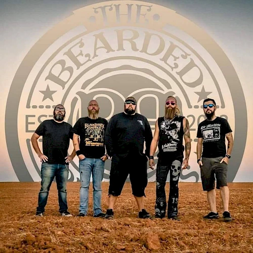 Five men with beards stand outdoors in front of a sign that says "The Bearded". They are wearing casual clothes and sunglasses, and standing on dirt.