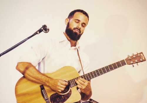 A man with a beard passionately playing an acoustic guitar, standing beside a microphone on a stand, indoors.