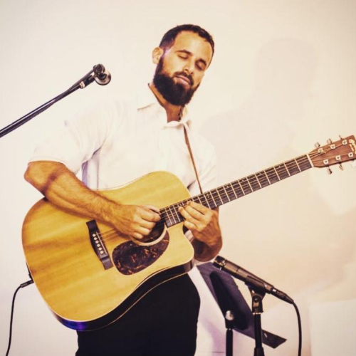 A man with a beard passionately playing an acoustic guitar, standing beside a microphone on a stand, indoors.