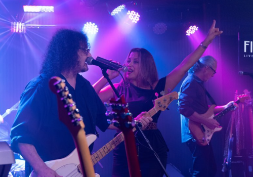 A live band is performing on stage under colorful lights, featuring a singer, bassist, and guitarist, creating an energetic atmosphere.