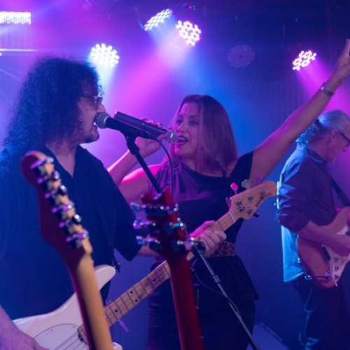 A live band is performing on stage under colorful lights, featuring a singer, bassist, and guitarist, creating an energetic atmosphere.