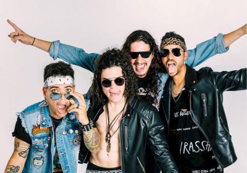 Four men are posing excitedly in rock-style outfits, wearing sunglasses and various accessories, against a plain white background.