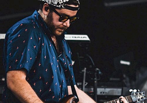 A person wearing sunglasses and a cap, playing an electric guitar on stage, with musical equipment in the background.