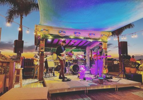 A small band performs on an outdoor stage at sunset, surrounded by palm trees and string lights, creating a relaxed and festive atmosphere.