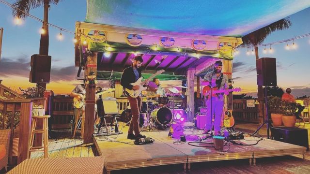 A small band performs on an outdoor stage at sunset, surrounded by palm trees and string lights, creating a relaxed and festive atmosphere.