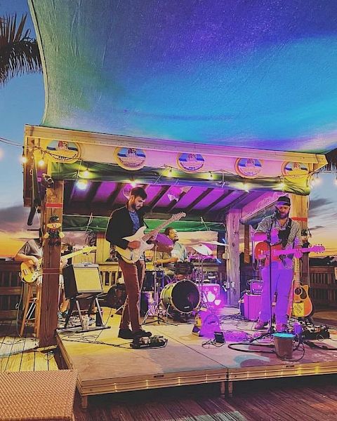 Two musicians perform on an outdoor stage with colorful lighting, under a canopy at sunset, surrounded by palm trees and string lights.