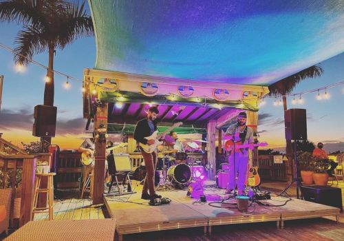 Two musicians perform on an outdoor stage with colorful lighting, under a canopy at sunset, surrounded by palm trees and string lights.