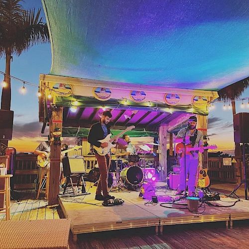 Two musicians perform on an outdoor stage with colorful lighting, under a canopy at sunset, surrounded by palm trees and string lights.