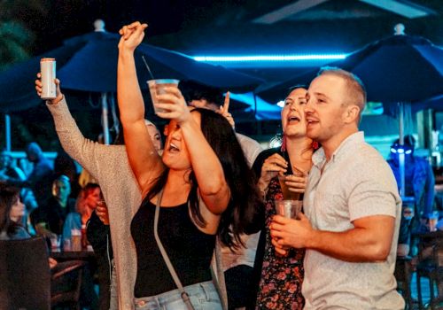 A group of people are enjoying a night out, holding drinks and dancing enthusiastically in a lively outdoor setting.