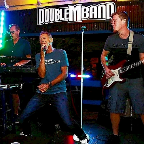 A band performing live on stage with a keyboardist, a lead singer, and a guitarist under a sign that reads "DoubleM Band".