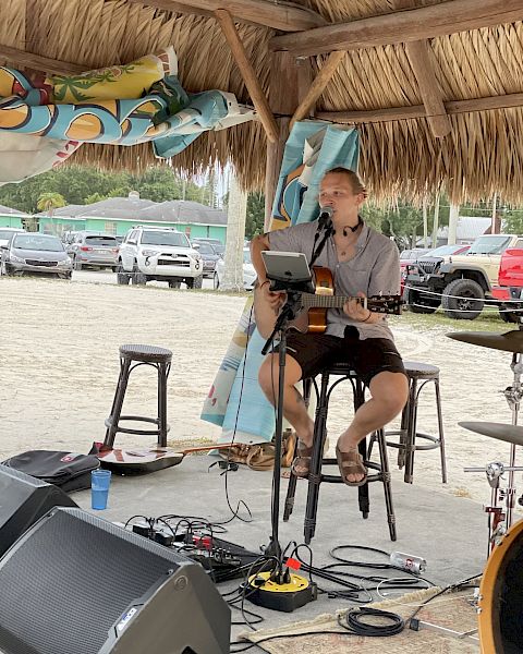 A person is playing guitar and singing into a microphone under a thatched roof, with music equipment around and a parking area in the background.