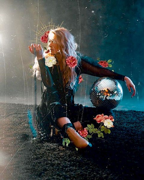 A person sits on the ground holding flowers, with a disco ball nearby. The background is dark and ethereal, with a mystical and artistic atmosphere.