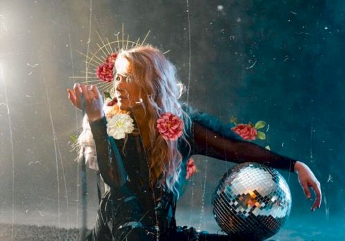 A person sits on the ground holding flowers, with a disco ball nearby. The background is dark and ethereal, with a mystical and artistic atmosphere.