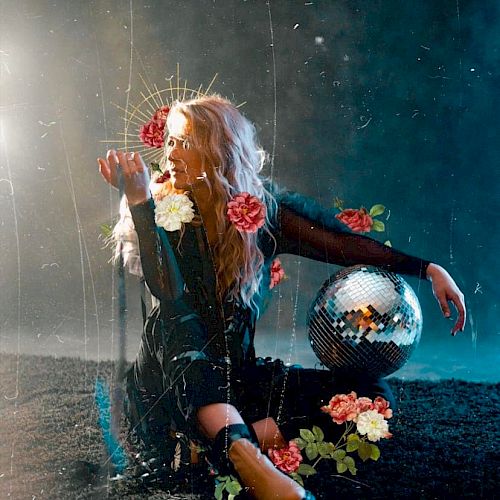 A person sits on the ground holding flowers, with a disco ball nearby. The background is dark and ethereal, with a mystical and artistic atmosphere.