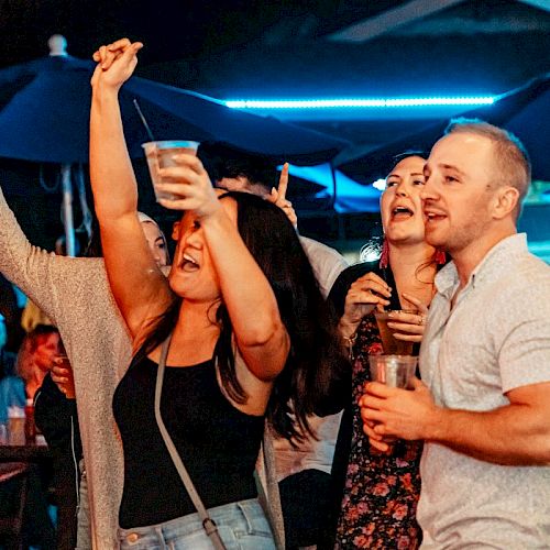 A group of people is happily celebrating at an outdoor venue, holding drinks and raising their hands, under the glow of blue lights.