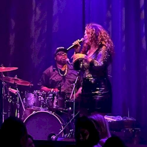 Two musicians perform on stage, a drummer and a singer with curly hair holding a microphone, under purple lighting, in front of an audience.