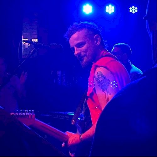 A musician is playing a guitar on stage under blue lights, wearing a sleeveless vest with tattoos visible on his arm.